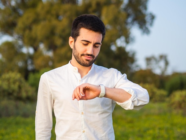 Photo young attractive spanish male checking the time on the wristwatch while posing in the park