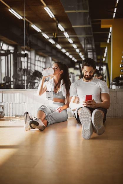 Young attractive sporty fitness couple sitting on the floor of the gym and making a break and relax time, leaning on the mirror with mobile and water.