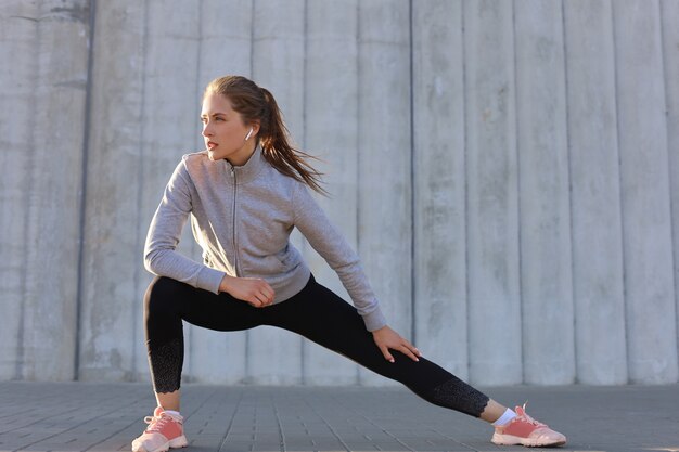 Young attractive sporty girl warming up outdoors, doing stretching at sunset or sunrise in city.