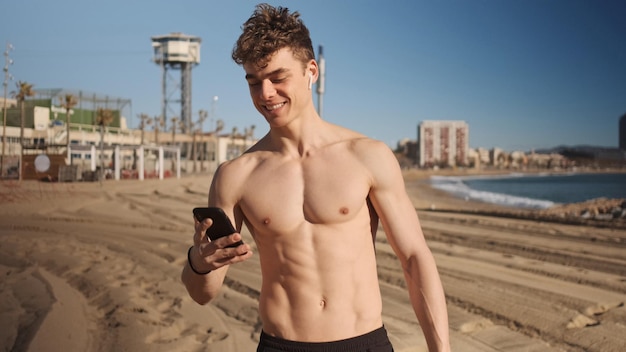 Young attractive sporty man with wireless earphones happily using smartphone during workout on city beach