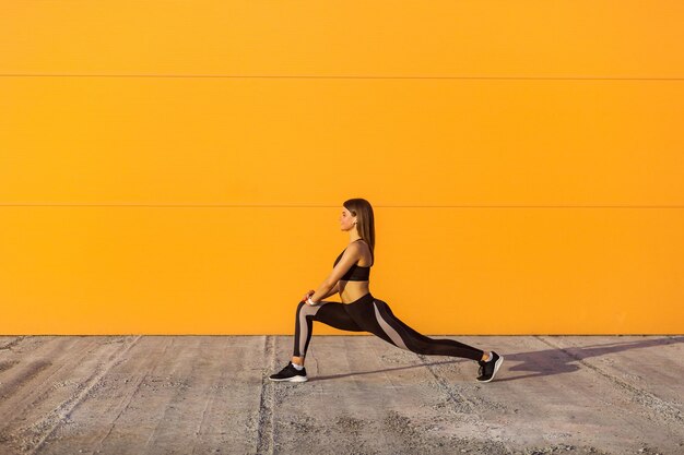 Young attractive sporty woman wearing black sporwear practicing sport exercises in morning on street, stretching legs and standing on ashtanga pose, orange wall background, outdoor