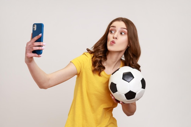 Young attractive teenager girl wearing yellow casual style T-shirt making selfie, having video call or broadcasting livestream with ball in hand. Indoor studio shot isolated on gray background.