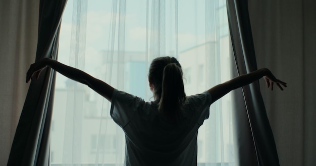 Young attractive woman approaches a window and opens curtains Warm morning light Hands pulling window curtain