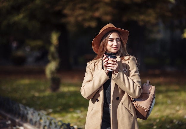 秋の服を着て、都市公園を歩きながらコーヒーを飲む若い魅力的な女性。