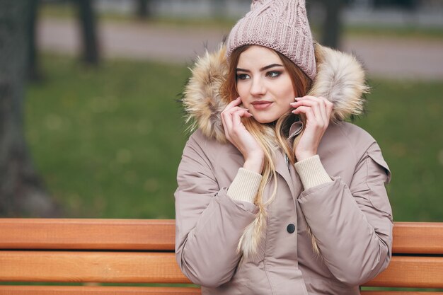 秋の服を着た若い魅力的な女性は、都市公園のベンチに座っています。女性は毛皮のスタイリッシュなジャケットを着ています。秋の時間。