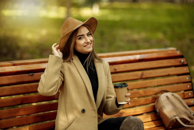 Young attractive woman in autumn clothes sits on bench and drinks coffee in city park. Woman dressed in stylish coat and felt hat. Autumn time