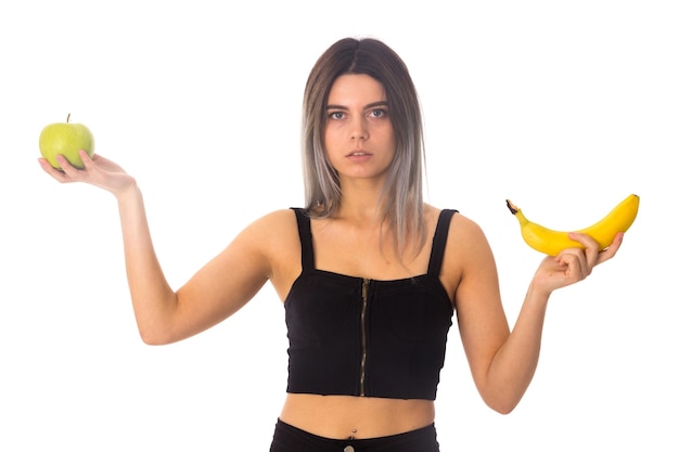 Young attractive woman in black top holding green apple and banana on white background in studio