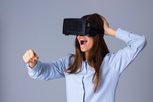 Young attractive woman in blue shirt using VR glasses and riding on grey background in studio