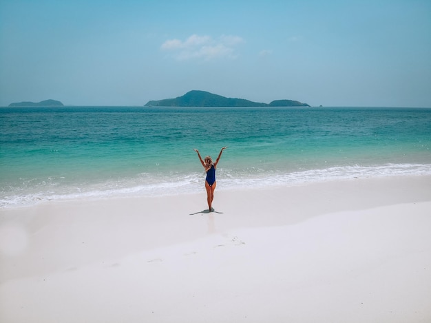 푸른 수영복을 입은 매력적인 젊은 여성이 모래 해변에 서서 일광욕을 하고 있습니다. 여름 휴가 개념