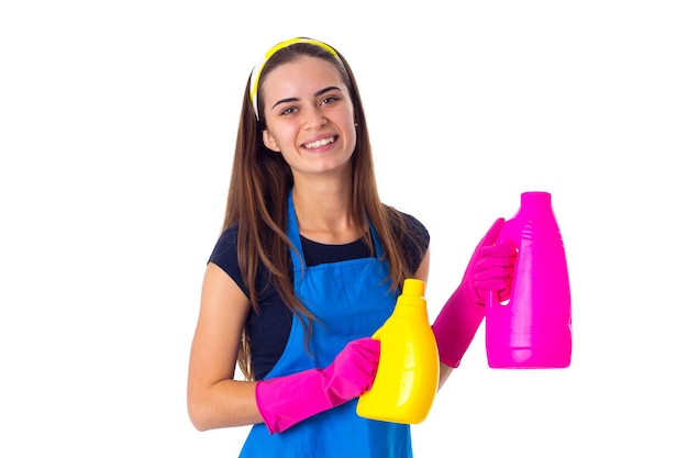 Young attractive woman in blue T-shirt and apron with pink gloves holding two detergents on white background in studio