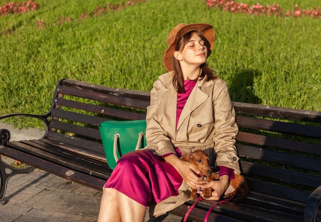 Young attractive woman dressed in autumn clothes with her lovely dachshund puppy sitting on a bench in the city