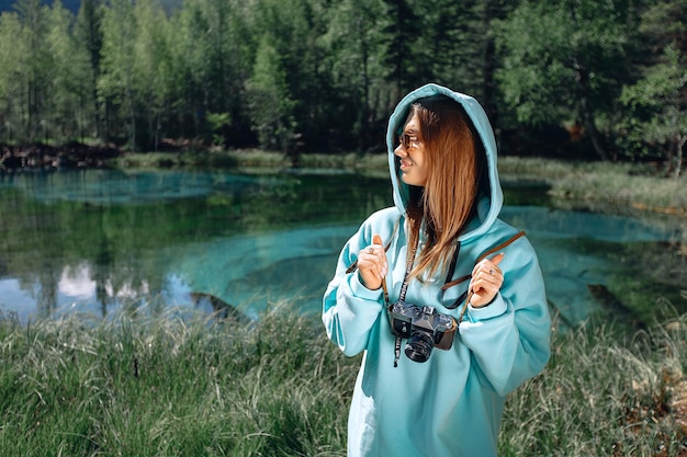 A young attractive woman dressed in a blue hoodie with a hood and sunglasses stands against the background of forest nature with grass, pond and trees holding a camera in her hands.