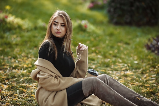세련된 코트, 터틀넥, 높은 스웨이드 부츠를 입은 젊은 매력적인 여자가 잔디에 앉아있다.