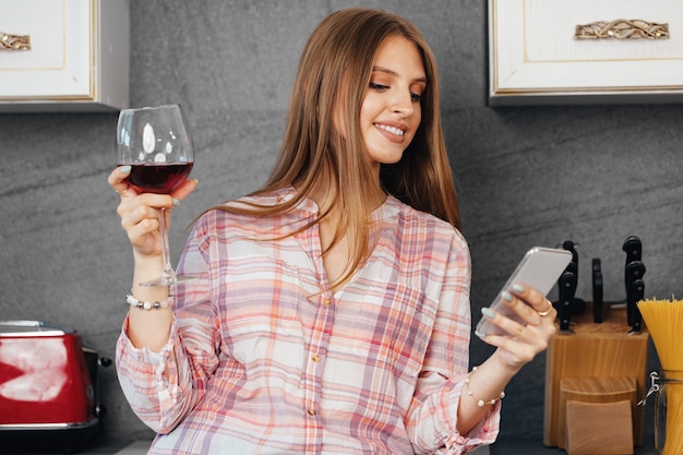 Фото Молодая привлекательная женщина пьет вино и использует смартфон на кухне, портрет