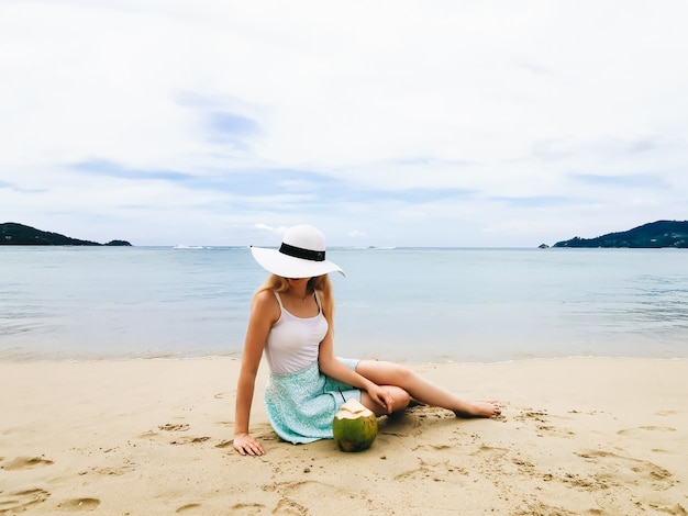 Young attractive woman in a hat on the shore of the blue sea sitting with a coconut, Thailand