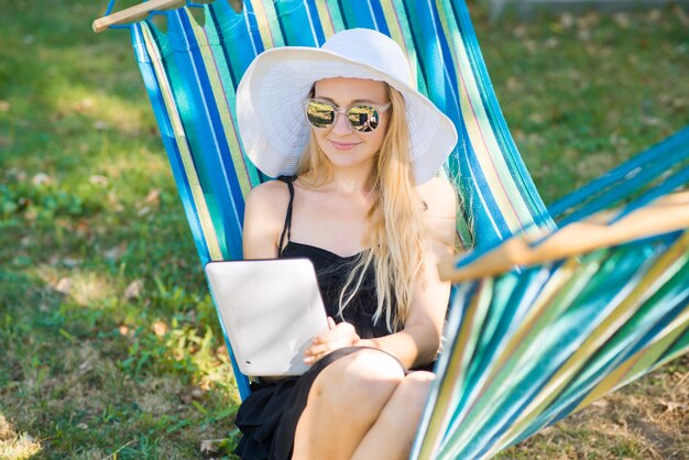 Young attractive woman in hat using a tablet on the hammock in the garden