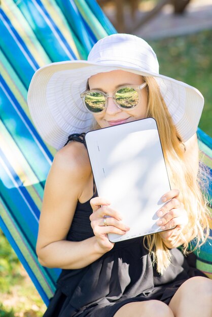 Young attractive woman in hat using a tablet on the hammock in the garden