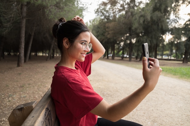 メガネと赤いシャツの20代の若い魅力的な女性は、ベンチに座って、携帯電話を持って、スペインの美しい公園で笑顔をしながら、selfieを取ります。彼女は電話を探しています