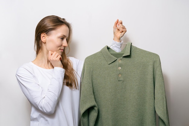 Молодая привлекательная женщина скептически смотрит на зеленый шерстяной свитер и выбирает наряд Концепция старых вещей и смена зимней одежды