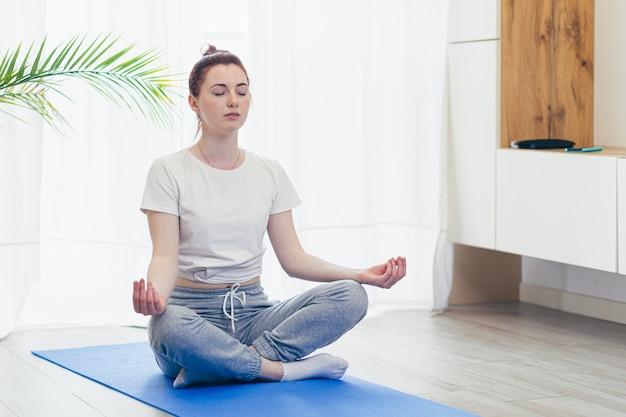 Young attractive woman is practicing yoga online using a mobile phone or smartphone app. Female yogi morning in casual clothes at home in living room, workout lotus position meditates on a mat