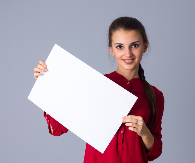 Young attractive woman in red blouse with plait showing white sheet of paper on grey background in studio