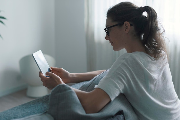 Young attractive woman relaxing at home in the living room she is sitting on the couch and connecting online with her digital tablet
