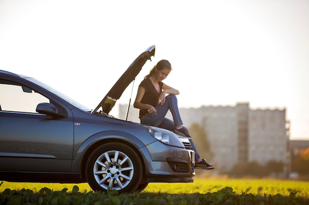 Giovane donna attraente che si siede sull'automobile con il cappuccio schioccato che controlla il livello dell'olio nel motore facendo uso dell'astina di livello sul fondo dello spazio della copia del cielo libero. trasporto, problemi dei veicoli e concetto di guasti.