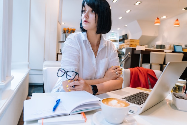 Young attractive woman sitting in office coworking and typing on her laptop keyboard computer and making notes in morning. Desk with a phone, notebook, glasses and a cup of coffee. Business concept