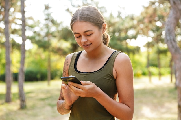  young attractive woman in sportswear using cellphone while working out in sunny green park