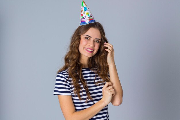 Young attractive woman in stripped T-shirt and celebration cap touching her long hair on grey background in studio