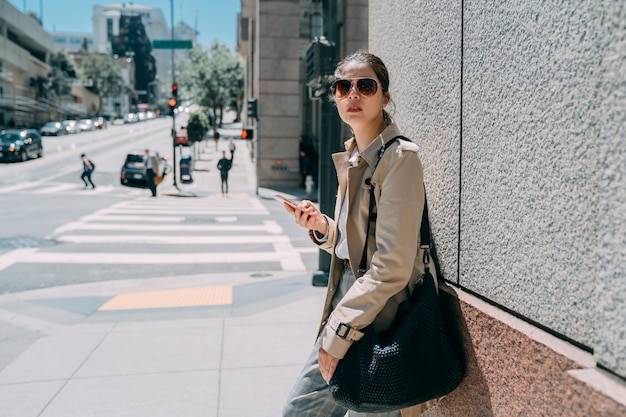 Young attractive woman in sunglasses stands outside on wall background. beautiful asian lady looking side and holding mobile phone. charming girl waiting friends on street lifestyle in summer time.