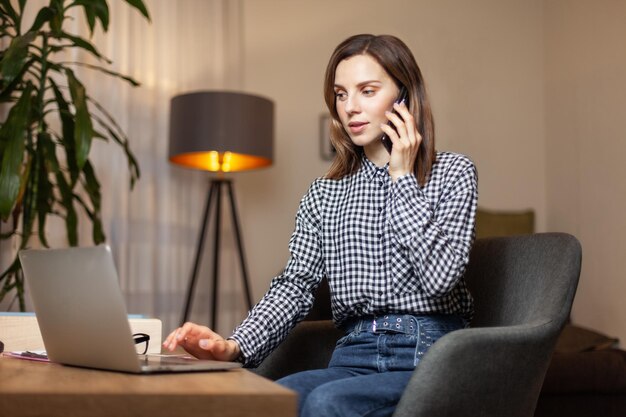 家のオフィスでラップトップを使って電話で話す魅力的な若い女性