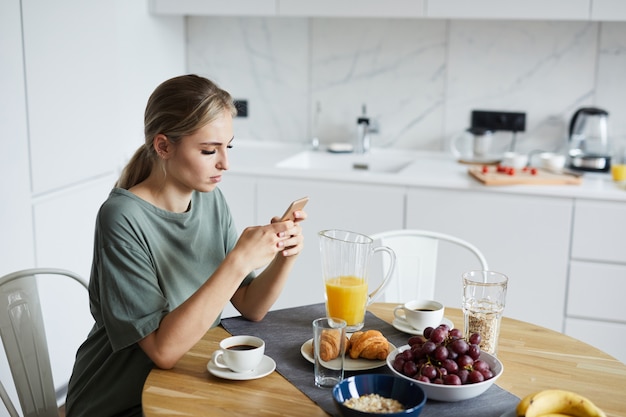 Young attractive woman using smartphone while having breakfast