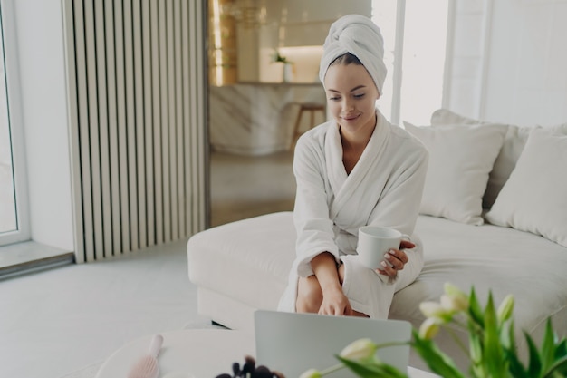 Молодая привлекательная женщина в белом халате и волосах, завернутых в полотенце, расслабляется на диване в гостиной после душа или ванны дома, счастливая женщина пьет чай или кофе, используя ноутбук и улыбается