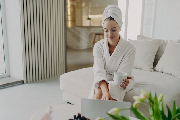 Young attractive woman in white bathrobe and hair wrapped in towel relaxing on sofa in living room after taking shower or bath at home, happy female drinking tea or coffee, using laptop and smiling