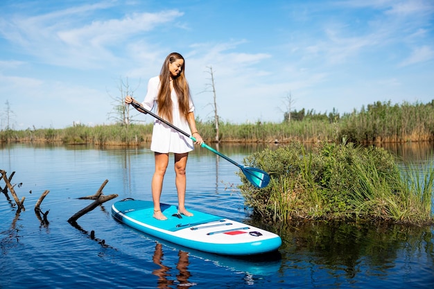 Young attractive woman in white dress standing on sup board in water holding paddle active weekend vacations wild nature outdoor