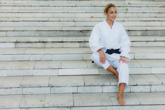 Young attractive woman in white kimono with black belt. Sport woman sitting on stairs outdoors. Martial arts