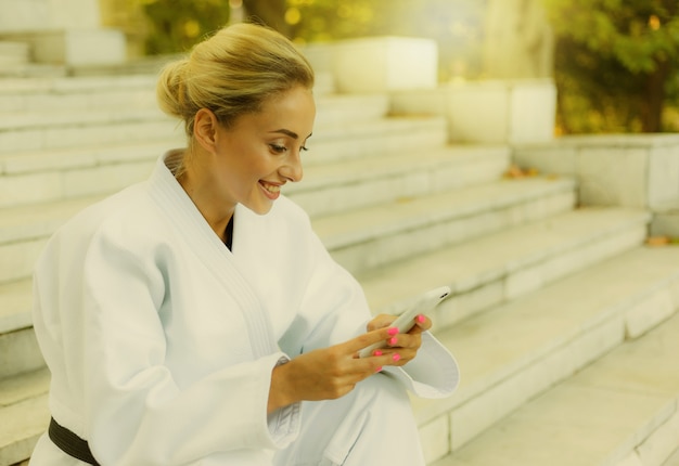 Young attractive woman in white kimono with black belt. Sport woman sitting on stairs and uses smartphone outdoors. Rest after training. Martial arts