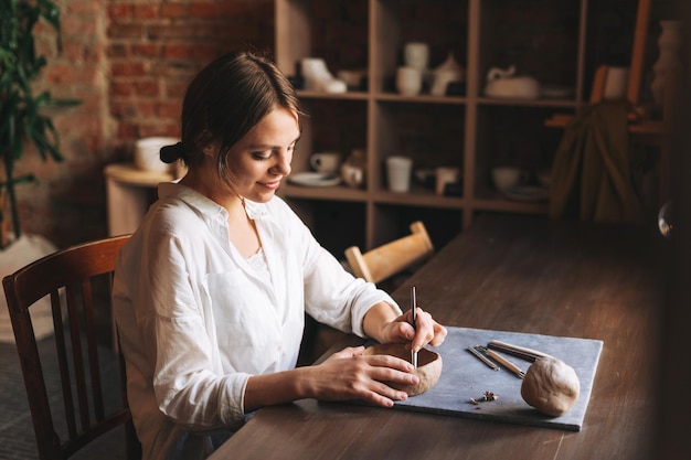 Young attractive woman in white shirt ceramic artist decorating clay plate with tool at table in pottery workshop Handmade work student freelance small business concept