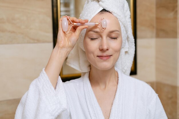 A young attractive woman with a bath towel on her head is\
massaging her forehead with a quartz gouache roller in the bathroom\
at home the concept of selfcare and cosmetic beauty procedures