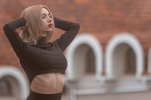 Young attractive woman with blonde hair wears black crop top and stands with hands behind her head Old city on background