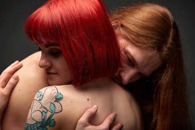 A young attractive woman with bright ruby hair tattoos and piercings is hugging a halfnaked man with