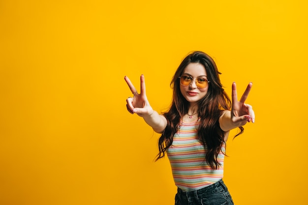 Young attractive woman with brunette hair in sunglasses standing and showing peace gesture