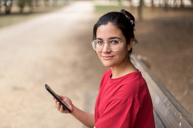 Giovane donna attraente con gli occhiali seduto su una panchina e usando il suo telefono in un parco, con una camicia rossa. siviglia, spagna