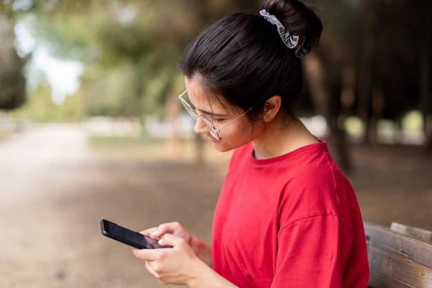 ベンチに座っていると、公園で彼女の電話を使用して、赤シャツのメガネの若い魅力的な女性。スペイン、セビリア