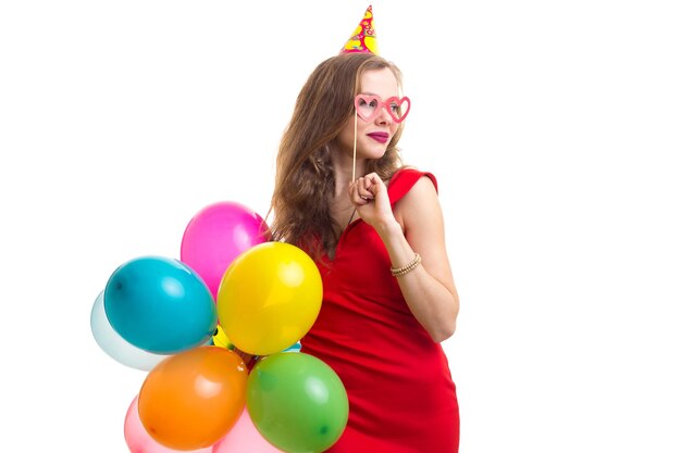 Young attractive woman with long chestnut hair in red dress with celebrating hat holding many colored balloons and card stick of glasses on white background in studio.