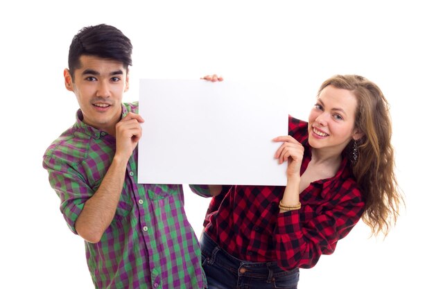 Young attractive woman with long chestnut hair and young pleasant man with dark hair in plaid shirts holding white placard on white background in studio