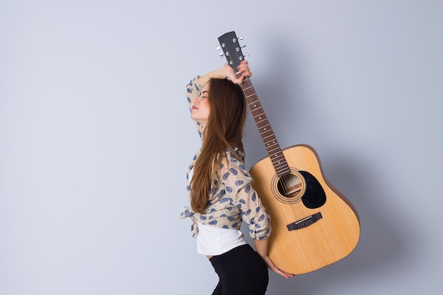 Young attractive woman with long hair in beige blouse and black trousers holding a guitar on gray background in studio