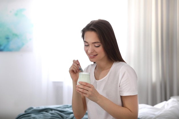 Young attractive woman with tasty yogurt on bed at home