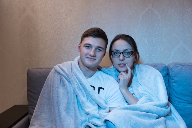 Молодая привлекательная пара, прижавшись друг к другу под одеялом на диване у себя дома, смотрит телевизор, вид спереди в синем сиянии от съемочной площадки